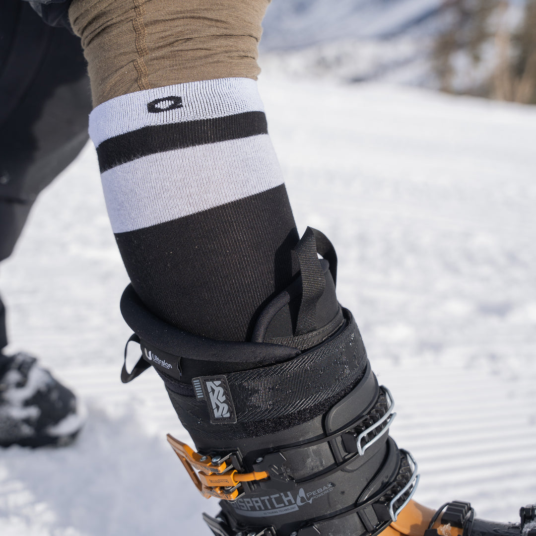 A close-up of a person’s leg wearing striped ski socks and a black ski boot with orange buckles, standing on snow with a blurred winter landscape in the background.