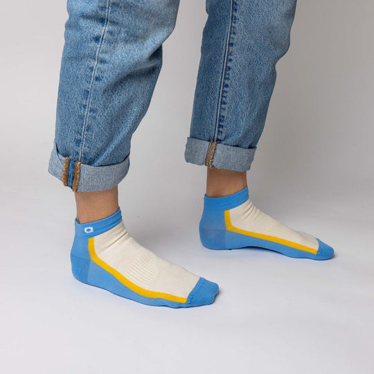 A person wearing rolled-up blue jeans and ankle socks with blue toes and heels, white tops, and yellow stripes, standing on a light-colored floor.