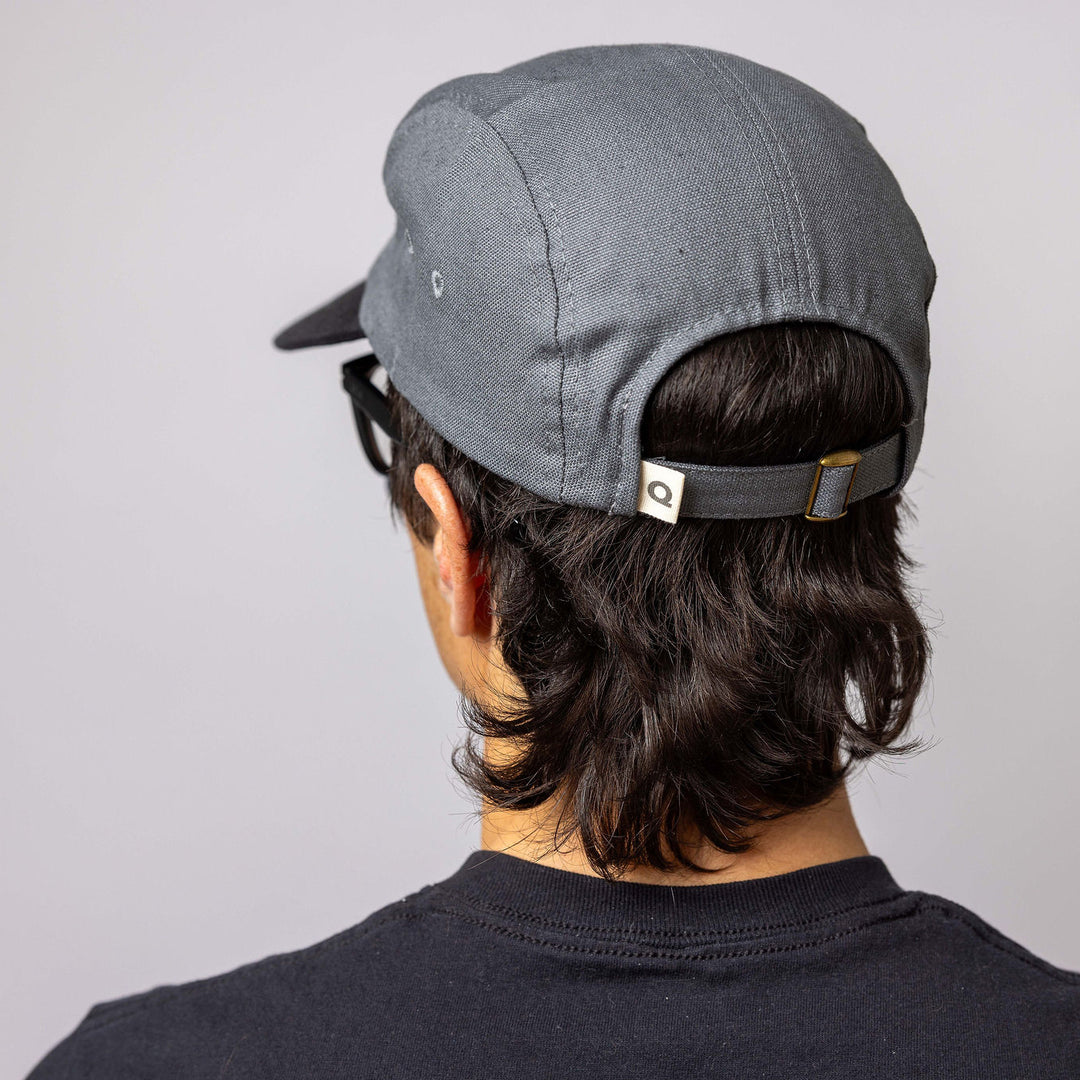A person with long dark hair wearing a black baseball cap backwards and a black t-shirt, shown from behind against a neutral background.