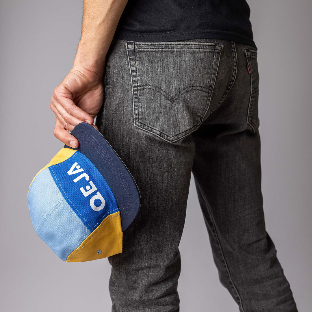 A person in black jeans holds a blue, yellow, and light blue cap with the word OEIA printed upside down on the brim, standing against a plain gray background.