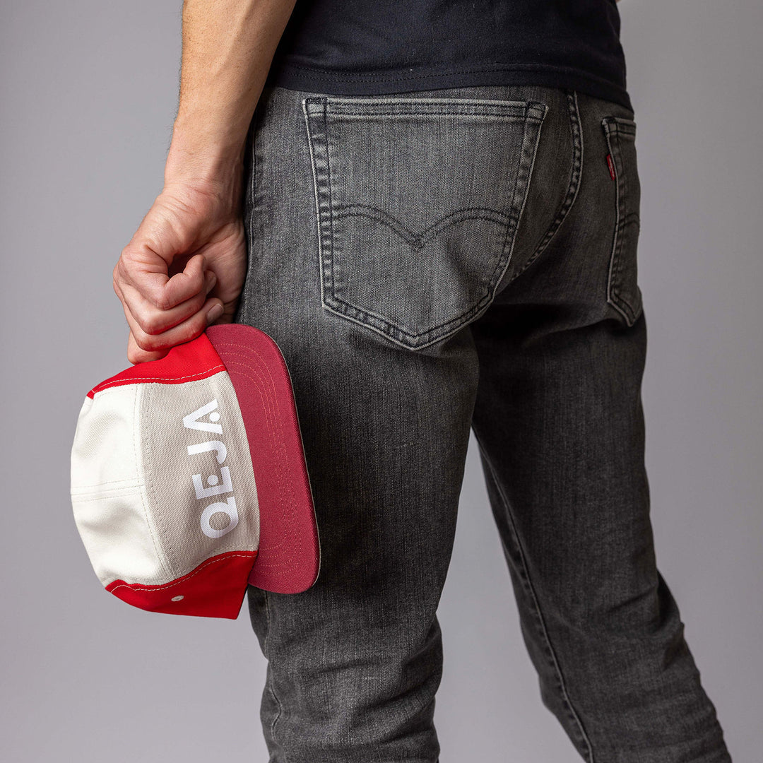 A person in grey jeans is standing with their back to the camera, holding a red and white cap with the word VEJA upside down in their left hand.