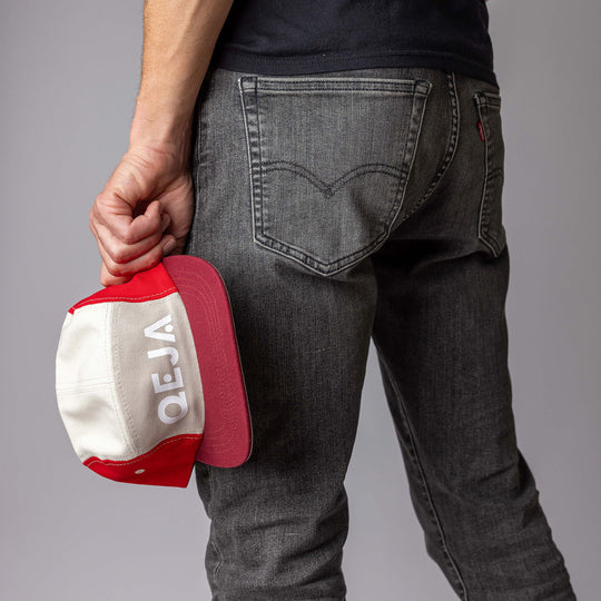 A person in grey jeans is standing with their back to the camera, holding a red and white cap with the word VEJA upside down in their left hand.