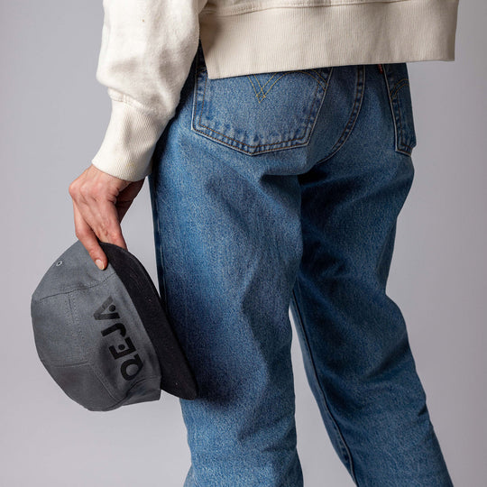 A person wearing blue jeans and a cream sweater holds a gray cap with VEJA written on it, standing against a plain gray background. The image is focused on the lower back and hand.