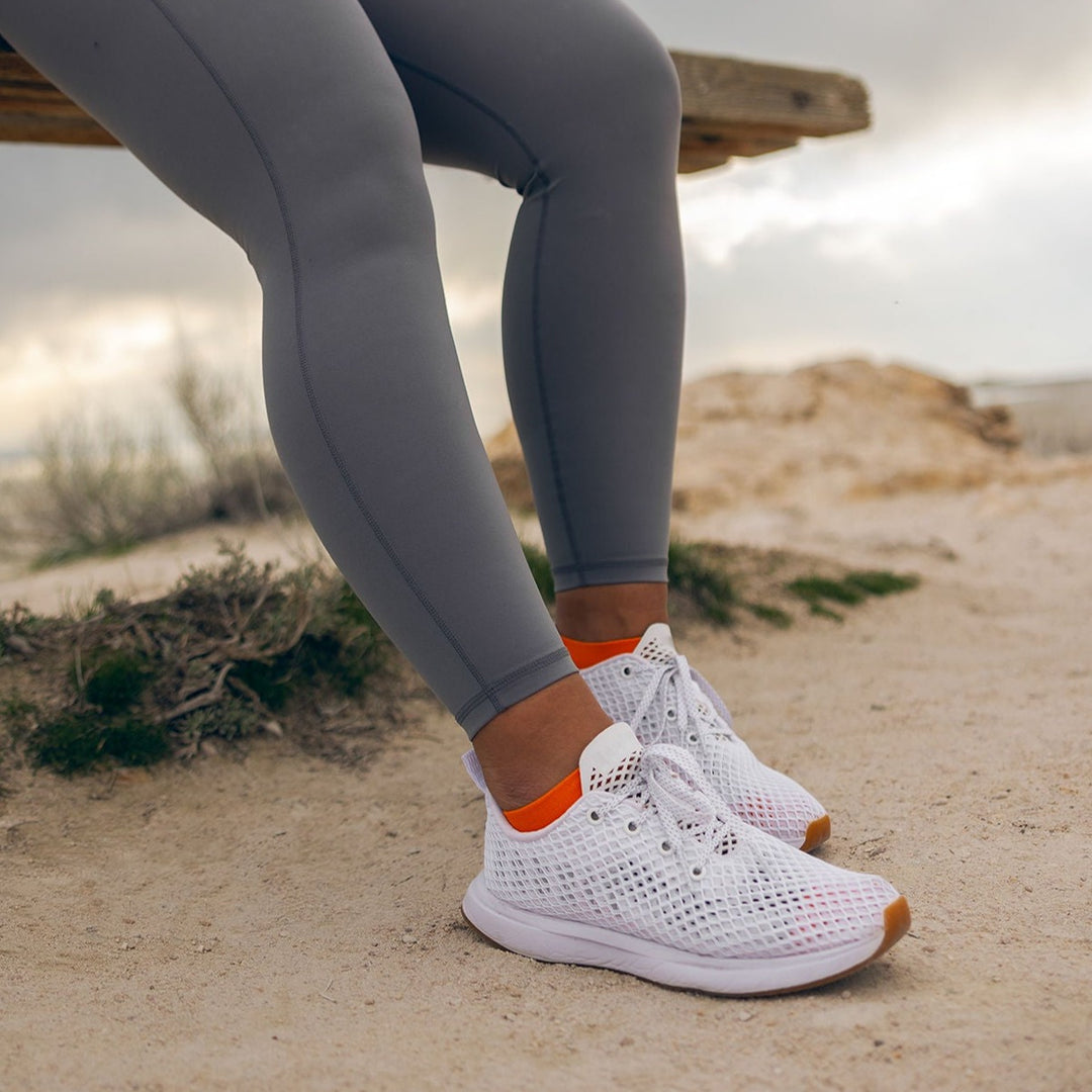 A person wearing grey leggings and white athletic shoes with orange accents sits on a wooden bench outdoors on a sandy surface.