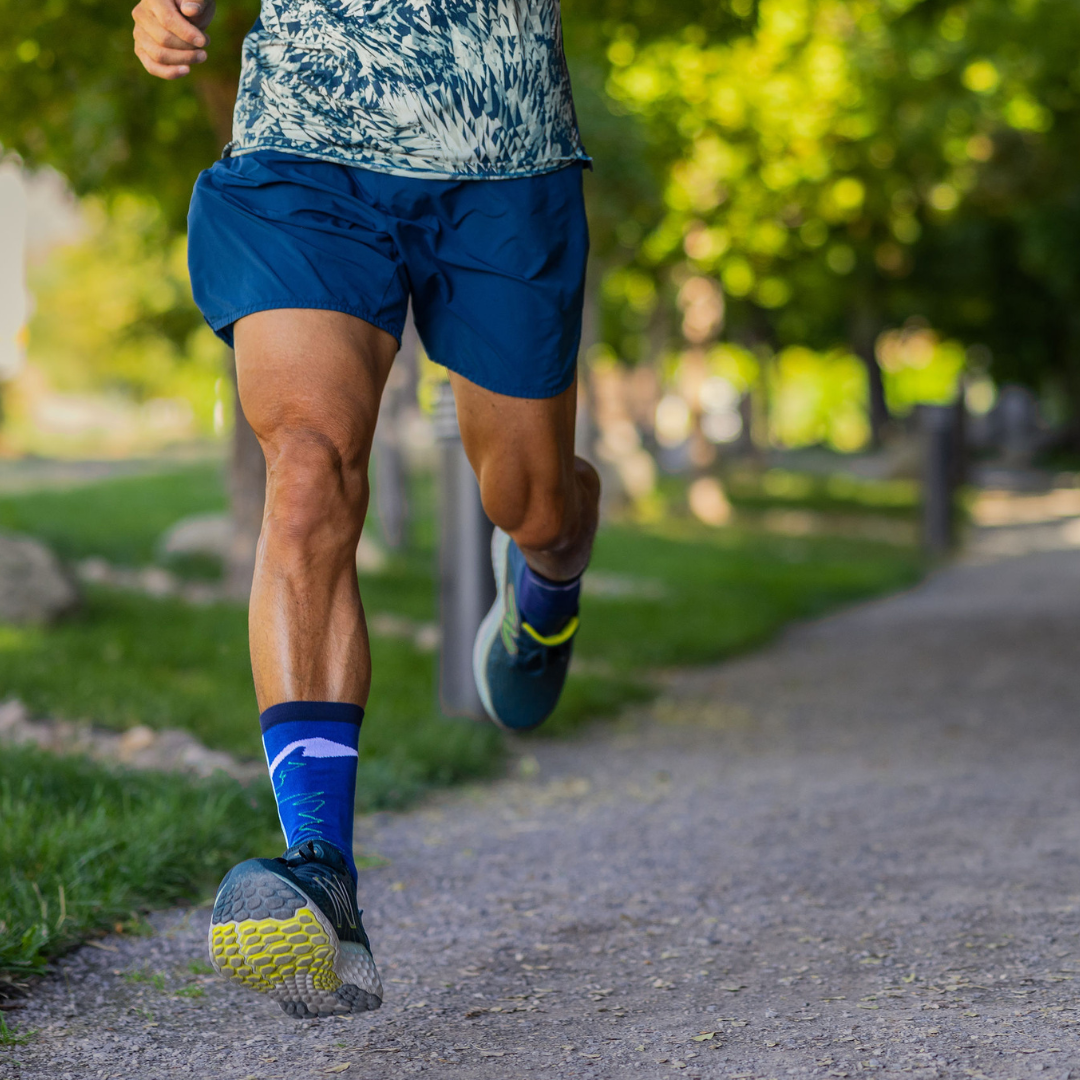 A person in blue shorts and athletic shoes is running on a gravel path in a park, with green grass and trees in the background. The image focuses on their legs in motion.