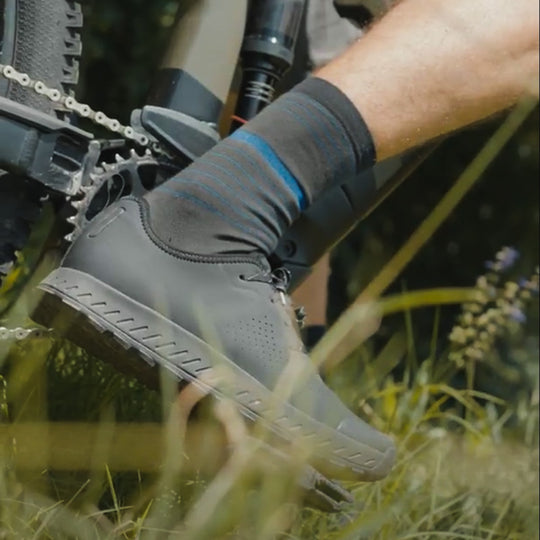 A close-up of a person’s foot on a bicycle pedal, wearing a black cycling shoe and striped socks, surrounded by green grass and plants.