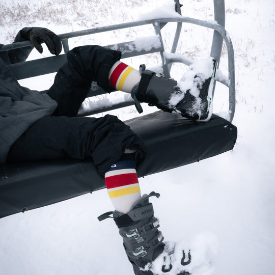 A person wearing ski boots and colorful striped socks sits on a snowy ski lift chair, with snow on their boots and the surrounding ground.