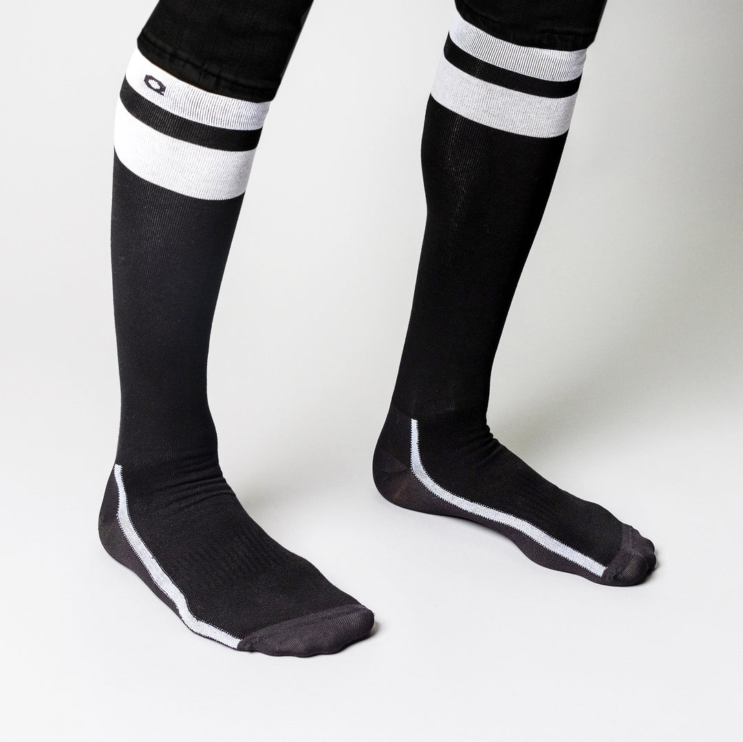Person wearing black knee-high socks with white stripes near the top, standing on a white background. Only the lower legs and feet are visible.